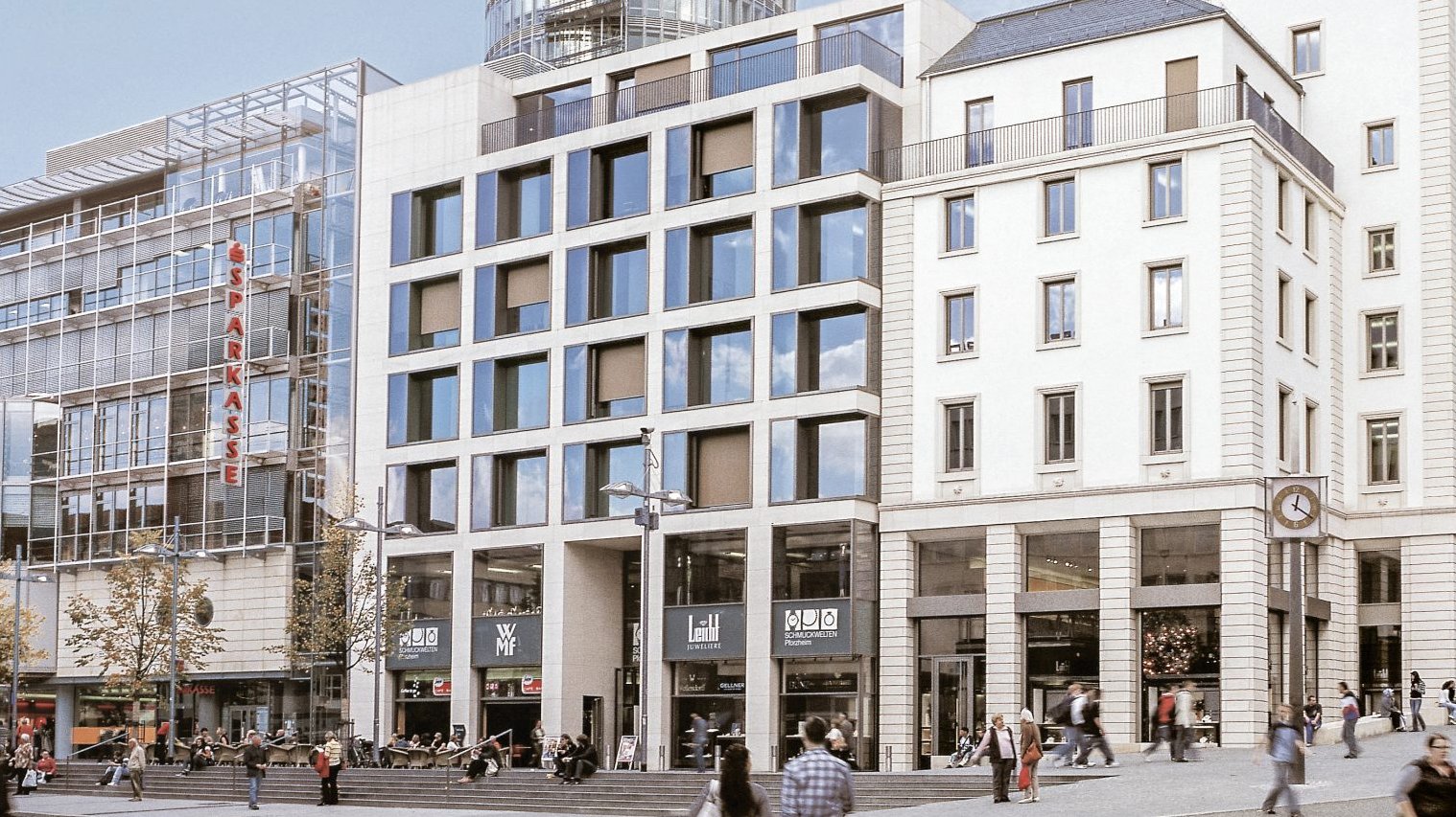 Moderne Stadtlandschaft mit zwei Gebäuden im Vordergrund. Menschen gehen über die Straßen und es gibt Cafés und Geschäfte im Erdgeschoss der Gebäude.