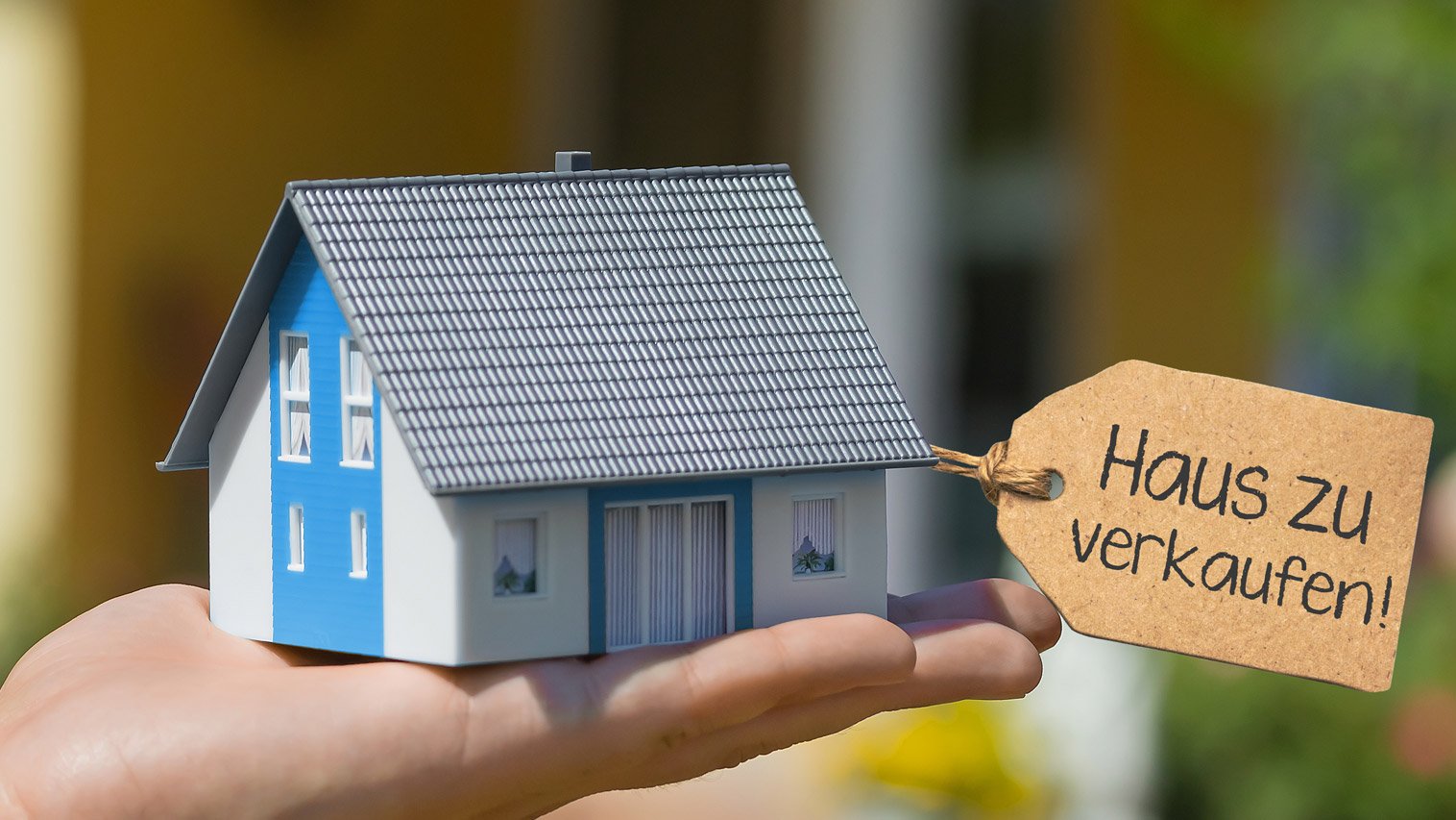 Eine Hand hält ein Miniaturmodell eines Hauses mit einem Schild, auf dem 'Haus zu verkaufen' steht.
