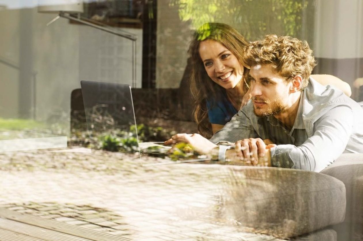 Ein Mann und eine Frau schauen gemeinsam lächelnd auf einen Laptop. Sie liegen auf einem Sofa.